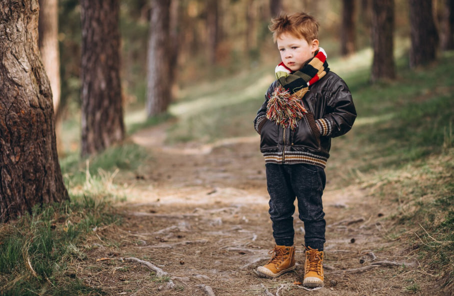 Young boy in a forest path, wearing a leather jacket, striped scarf, and tan boots, holding a colorful pompom.