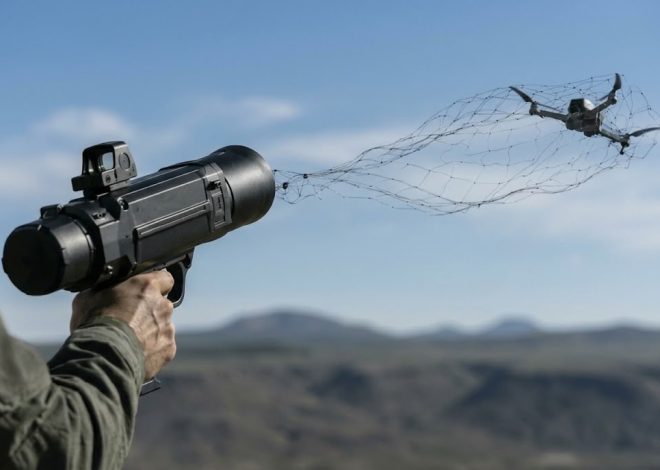 Person aiming a black net launcher and releasing a net toward a quadcopter drone flying low over a desert landscape