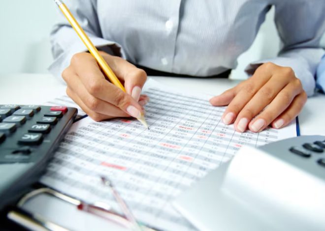 Person writes on a printed spreadsheet with a yellow pencil, calculator nearby, suggesting accounting work.