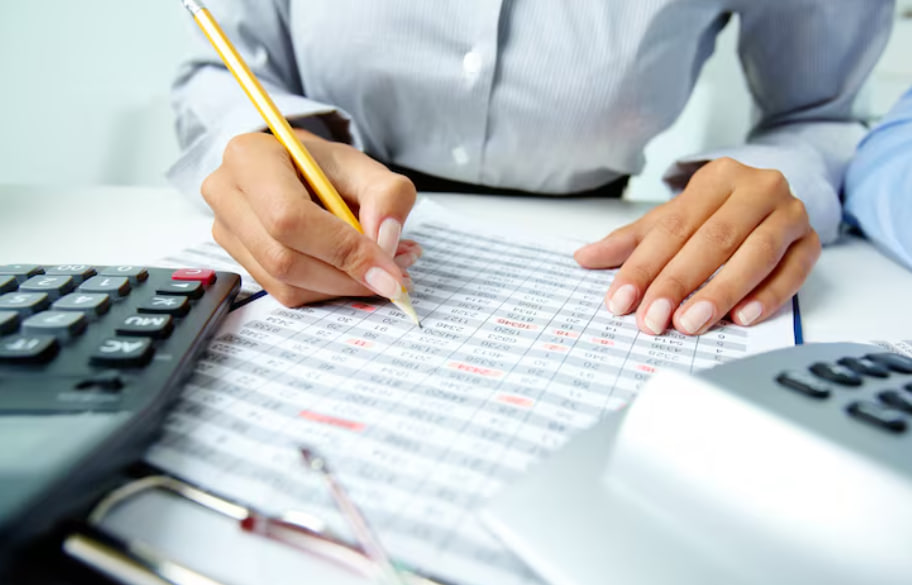 Person writes on a printed spreadsheet with a yellow pencil, calculator nearby, suggesting accounting work.