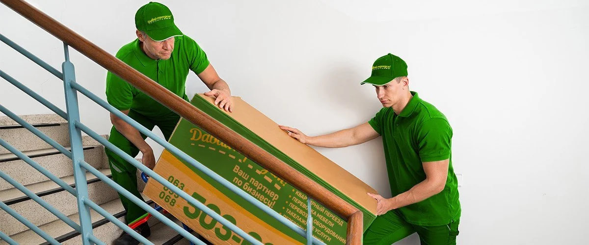 Two movers in green uniforms carry a large cardboard box down a stairwell, working together to move it carefully down the stairs.