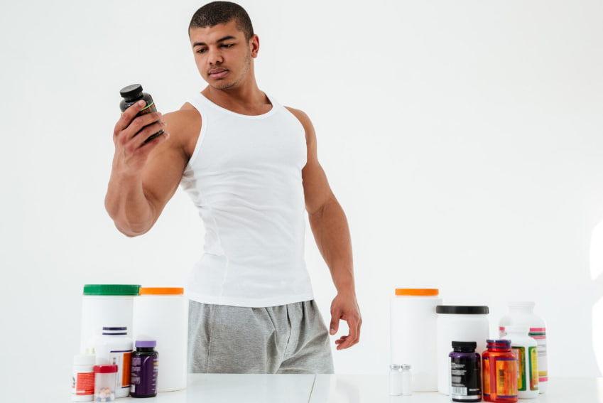 Fit man in a white tank top holds a supplement bottle while standing behind a table with protein jars and pill bottles on display.