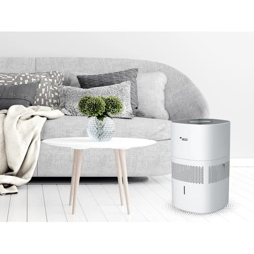 Modern living room with a grey sofa, cushions, cream throw, round white coffee table, vase with green plants, and a white cylindrical air purifier on the floor.