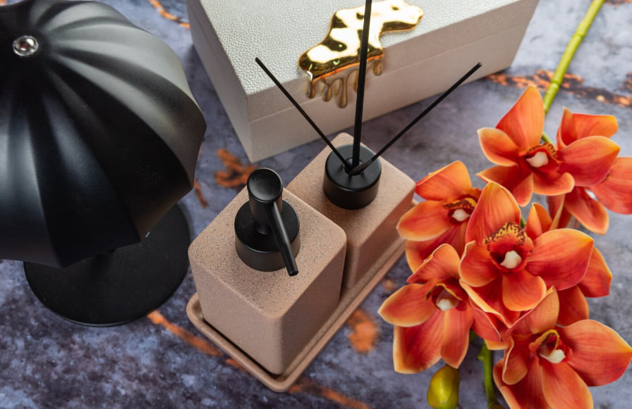 Reed diffuser arrangement with black bottles on beige blocks beside bright orange flowers on a dark stone surface.