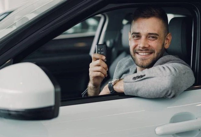 Man smiling in the driver's seat of a white car, holding up a key fob toward the camera.