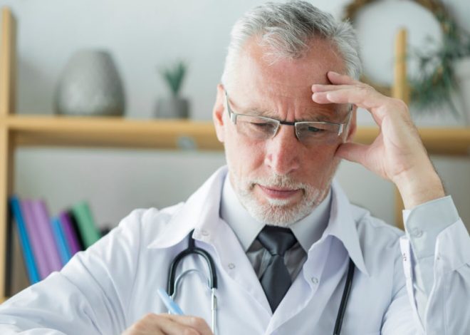 Older male doctor in a white coat and glasses, resting his head on his hand in a thoughtful or stressed pose.