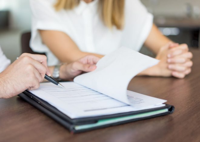 Person signs documents with a pen over a binder on a wooden table while another person sits nearby in a meeting setting.