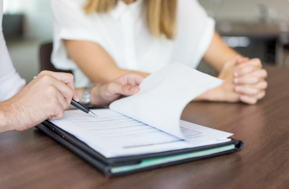 Person signs documents with a pen over a binder on a wooden table while another person sits nearby in a meeting setting.
