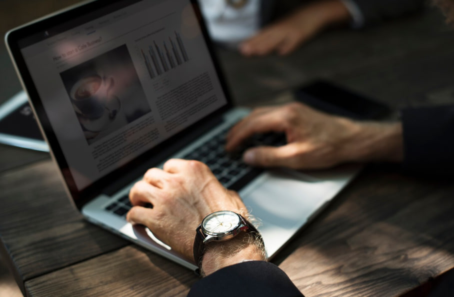 Hands typing on a laptop with a wristwatch visible; screen shows a news article and bar chart.÷