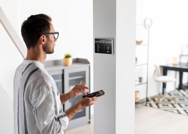 Man in a striped shirt uses a remote to control a wall-mounted smart home panel in a bright open living area.