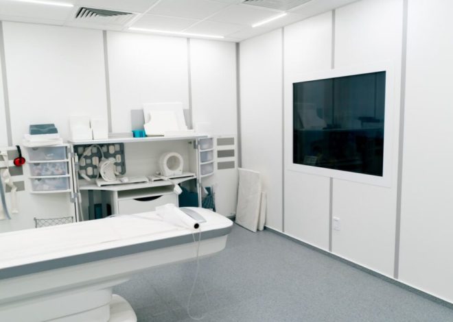 Empty radiology suite with a patient table and imaging equipment, wall-mounted monitor on the right.