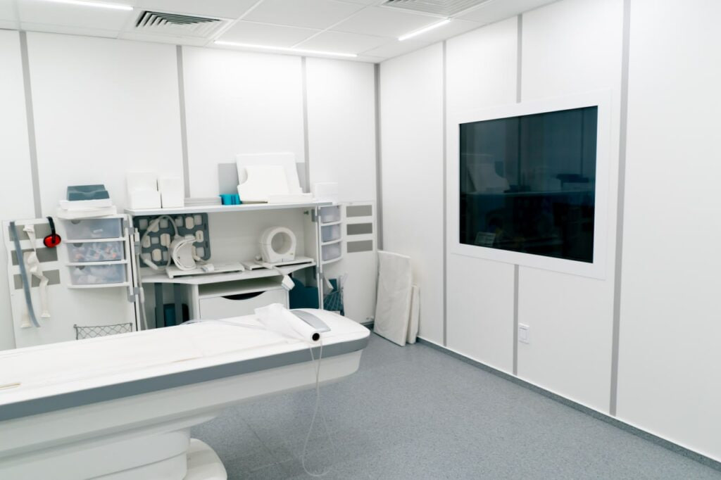 Empty radiology suite with a patient table and imaging equipment, wall-mounted monitor on the right.