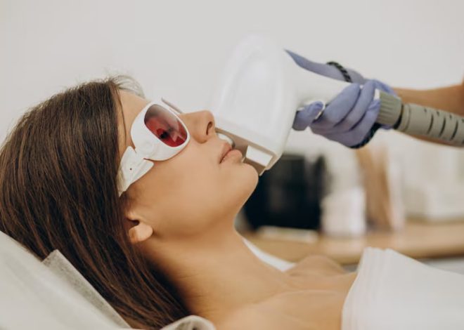 Woman undergoing a cosmetic procedure: wearing protective goggles while a clinician uses a handheld device near her mouth/cheek with a gloved hand in a clinical setting.