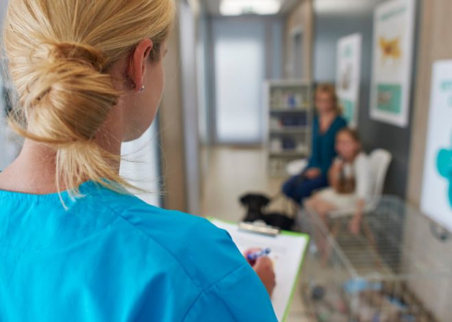 Healthcare professional in blue scrubs notes on a clipboard in a clinic lobby as patients sit in the background.