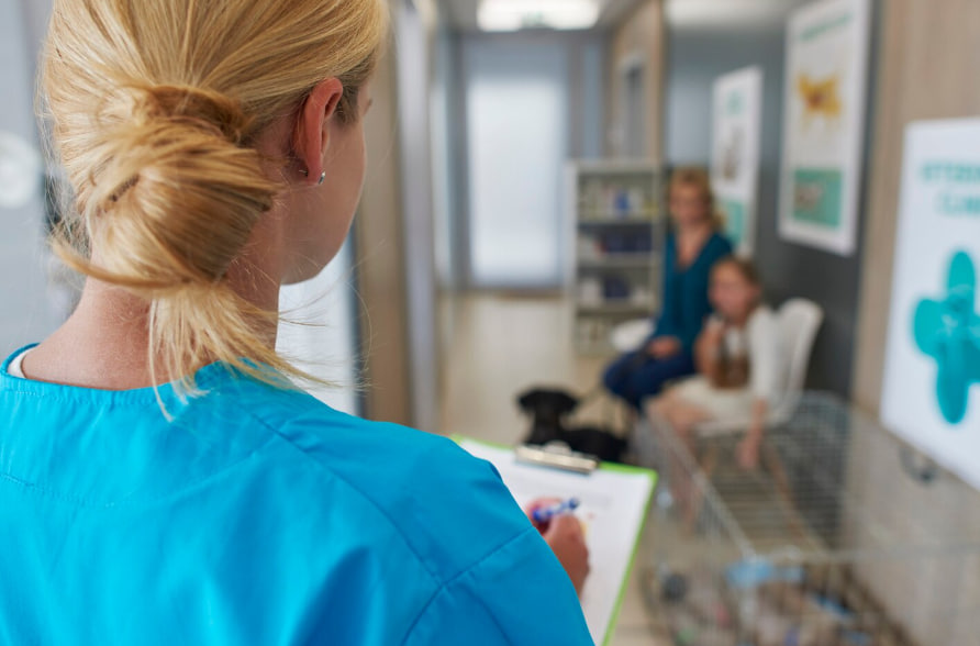 Healthcare professional in blue scrubs notes on a clipboard in a clinic lobby as patients sit in the background.