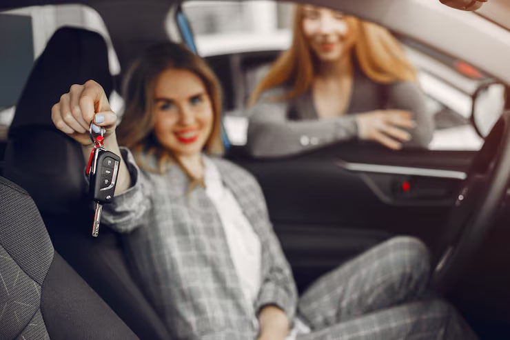 Woman in the driver's seat holds up car keys toward the camera as another woman smiles in the passenger seat behind her.