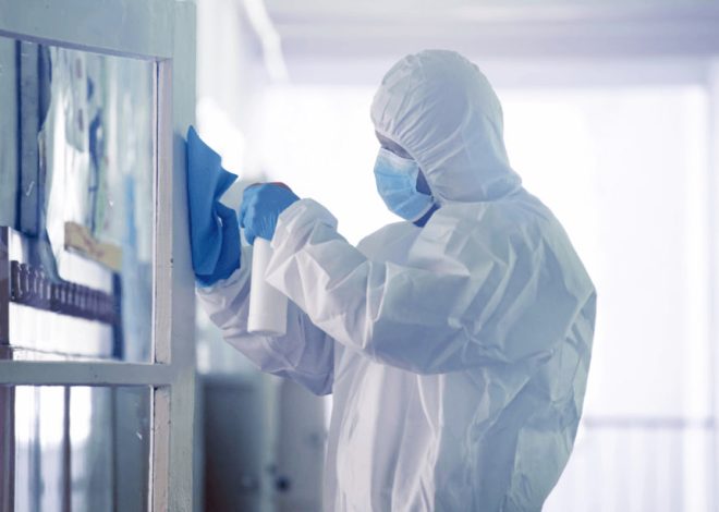 Person in full PPE wipes a glass door with a blue cloth in a clean, sterile corridor or lab setting.
