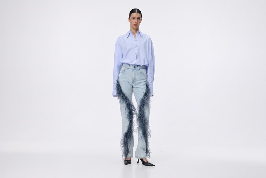 Model wearing a light blue button-down shirt tucked into high-waisted, light-wash jeans with blue feather fringe along the legs, black heels, on a white studio background.