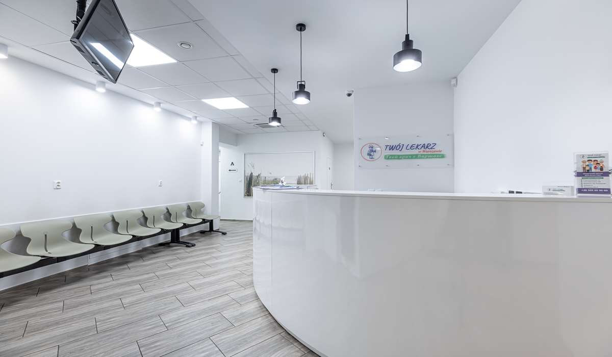 Modern medical clinic reception with a curved white front desk, pale green chairs along the wall, and a banner reading 'Twój Lekarz' in the background; bright, clean space.