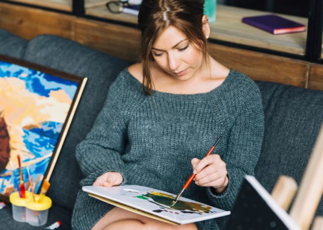Woman in a gray knit sweater sits on a sofa and paints with a brush on a sketchpad, with art supplies nearby.