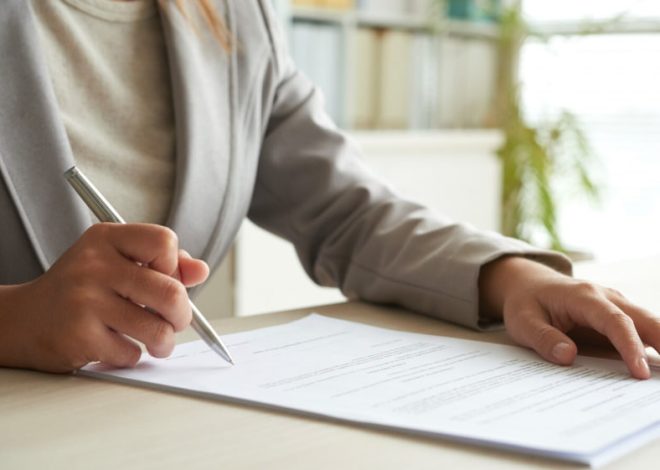 Person in a blazer signing a document with a silver pen at a desk (likely a contract)