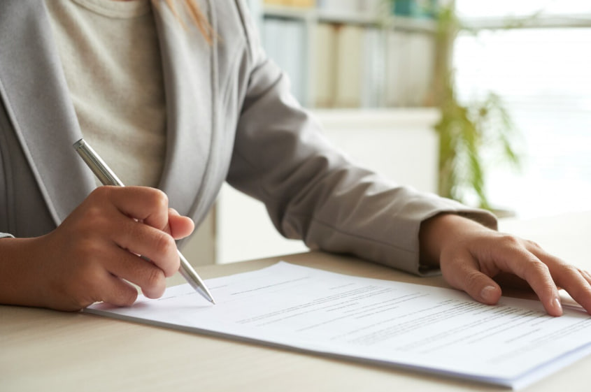 Person in a blazer signing a document with a silver pen at a desk (likely a contract)