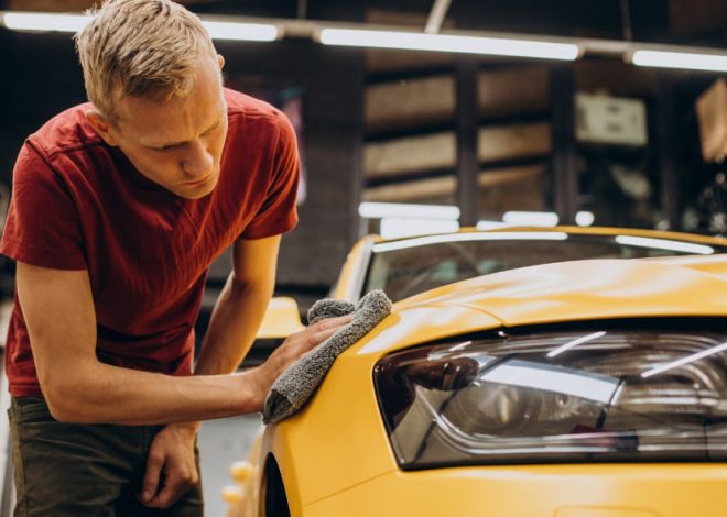 Man in a red shirt polishing the hood of a yellow car with a gray microfiber cloth in a workshop setting.