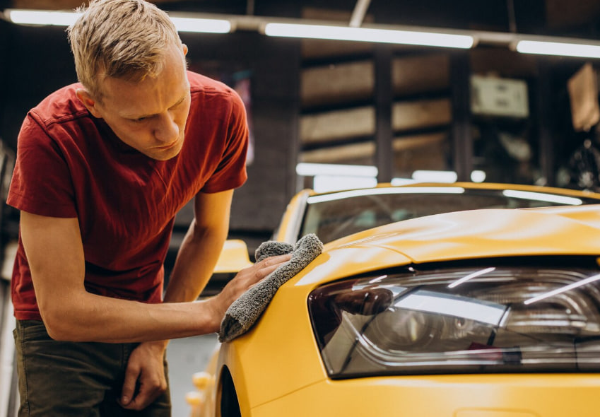 Man in a red shirt polishing the hood of a yellow car with a gray microfiber cloth in a workshop setting.