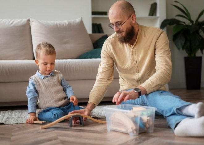 Father and toddler play with a wooden train set on the living room floor, near a white sofa.