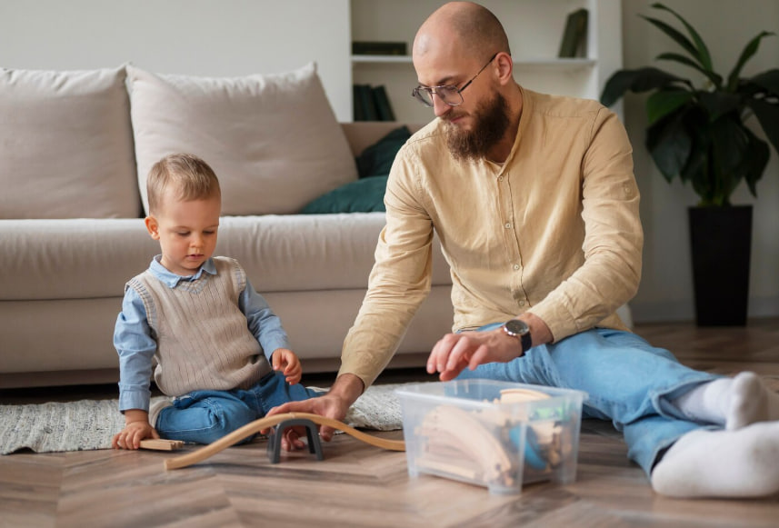 Father and toddler play with a wooden train set on the living room floor, near a white sofa.