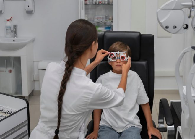 Optometrist adjusts trial lenses on a young patient wearing red-rimmed glasses during an eye exam in a clinicroom.