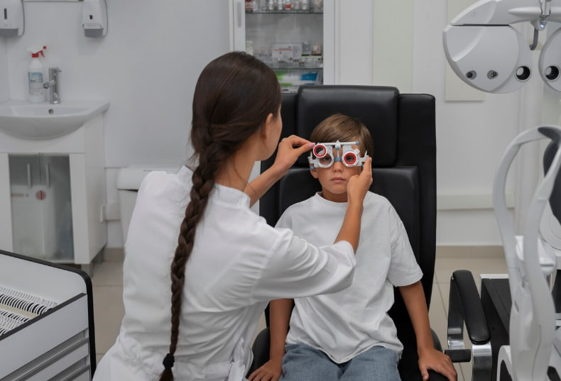 Optometrist adjusts trial lenses on a young patient wearing red-rimmed glasses during an eye exam in a clinicroom.