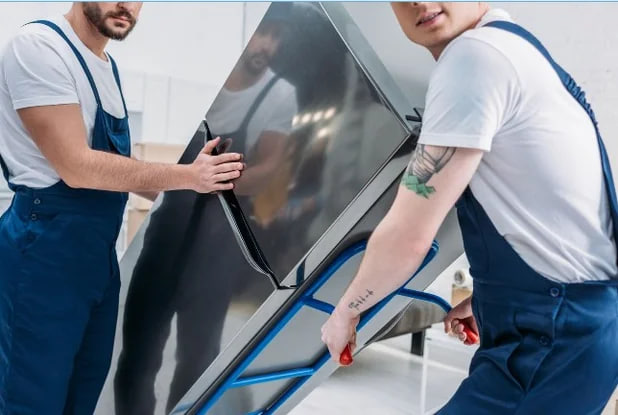 Two movers in blue overalls transport a large stainless-steel refrigerator on a dolly in a bright room.