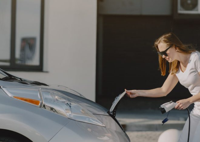 Woman in a white shirt plugs in an electric car’s charging cable beside the front hood.