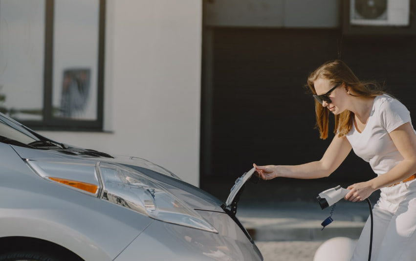 Woman in a white shirt plugs in an electric car’s charging cable beside the front hood.