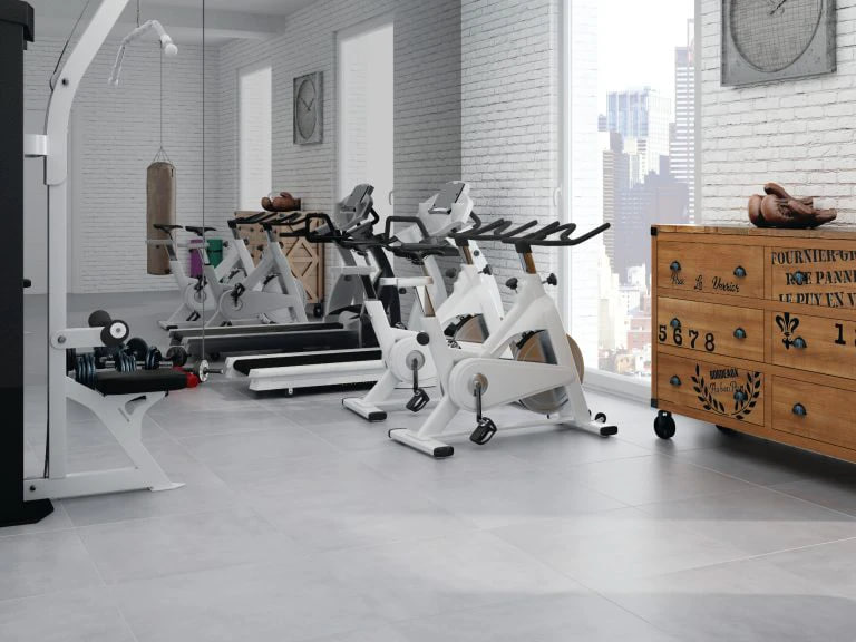 Row of white stationary bikes in a bright, modern gym by large windows with a wooden dresser nearby.