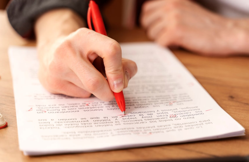 Person writing with a red pen on a printed document on a wooden table