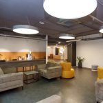 Modern lounge area with gray sofas, yellow chairs, and a coffee bar along a wooden counter.