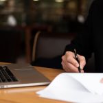Businessperson in a suit signs a document with a pen beside an open laptop on a wooden desk