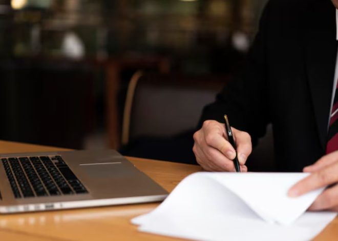 Businessperson in a suit signs a document with a pen beside an open laptop on a wooden desk
