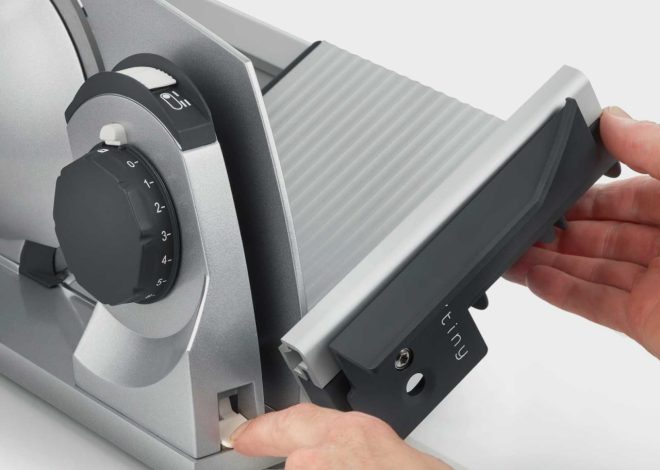 Close-up of a manual food slicer being adjusted: a person slides in a blade guard while turning a numbered adjustment dial.