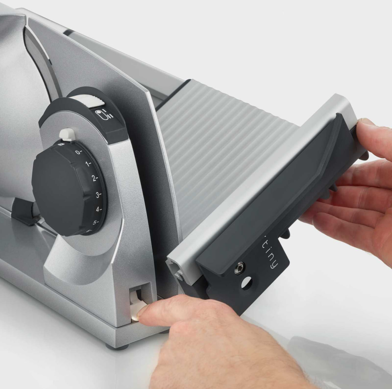 Close-up of a manual food slicer being adjusted: a person slides in a blade guard while turning a numbered adjustment dial.