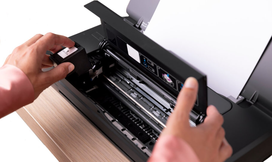 Person’s hands are installing or replacing a toner cartridge inside a black printer with paper loaded at the top (printer maintenance).