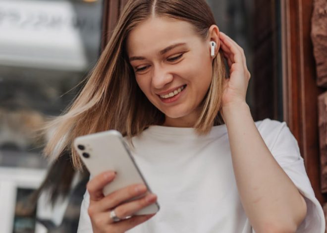 Smiling young woman wearing wireless earbuds looks at her smartphone outdoors by a brick building.