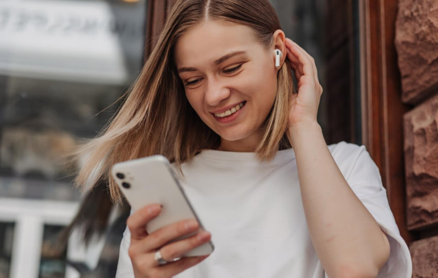 Smiling young woman wearing wireless earbuds looks at her smartphone outdoors by a brick building.