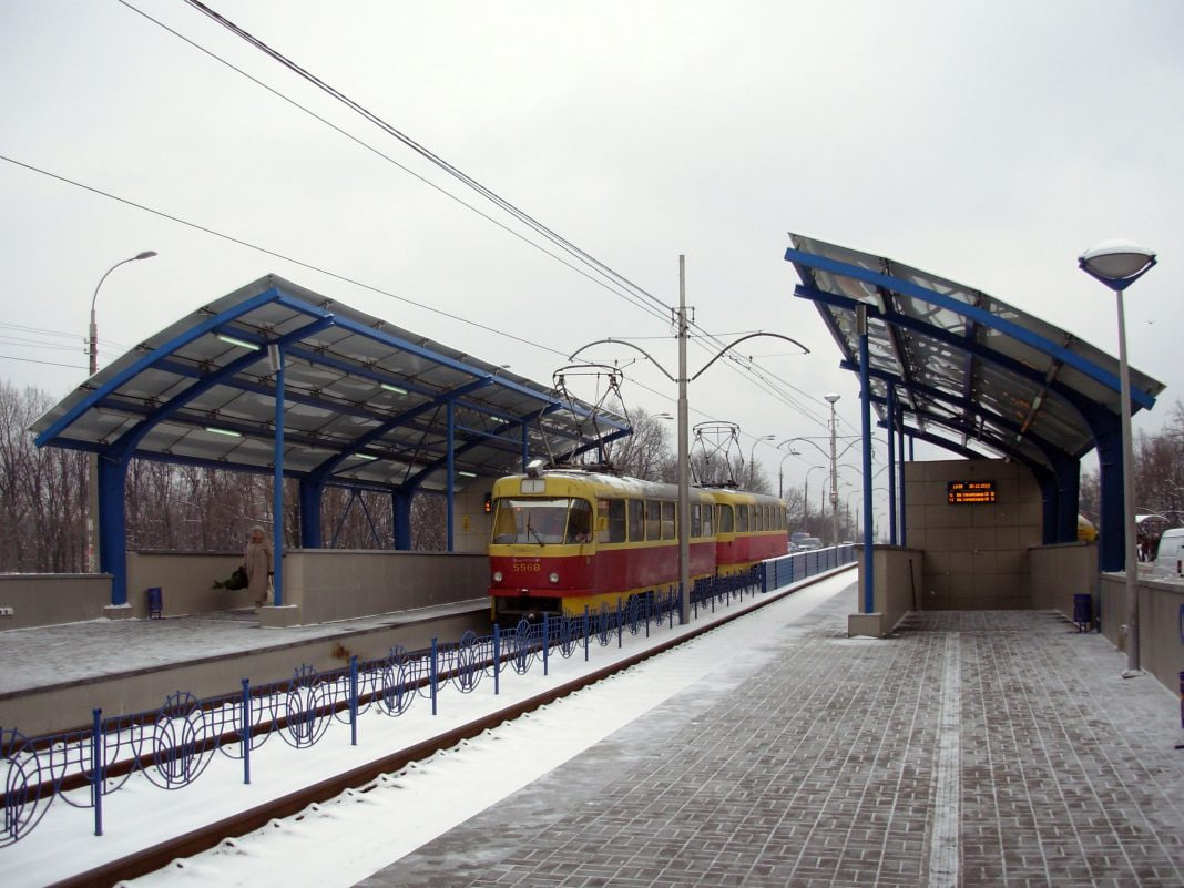 photo_2026-10-20_16-16-30 - TYME BLOG Red-and-yellow tram at a snow-covered station with blue curved canopies and overhead wires.