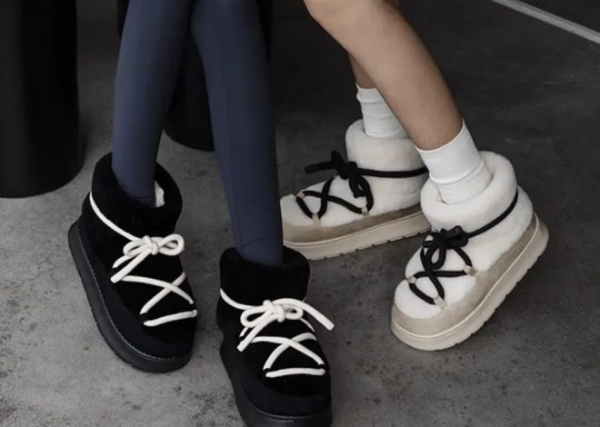 Two pairs of shearling-lined boots with chunky laces (black and white) placed on a concrete floor, worn with jeans and white socks on one side and bare ankles on the other.