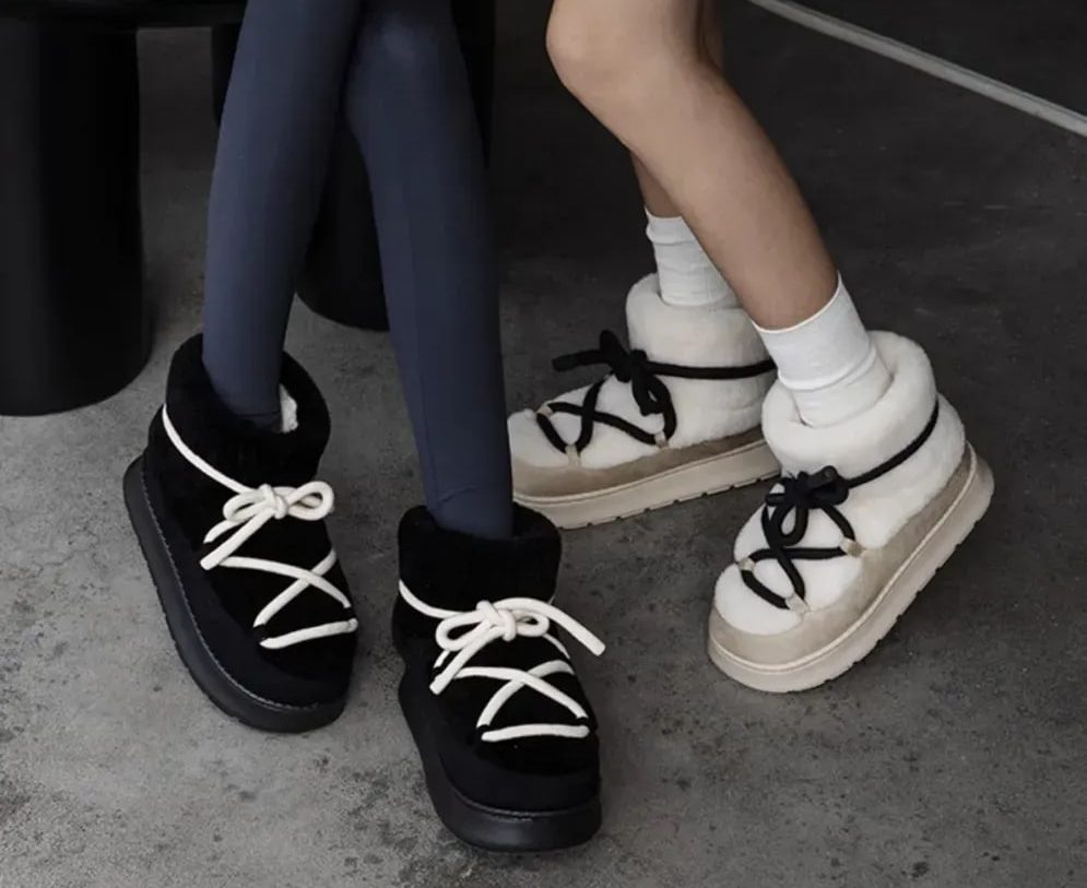 Two pairs of shearling-lined boots with chunky laces (black and white) placed on a concrete floor, worn with jeans and white socks on one side and bare ankles on the other.