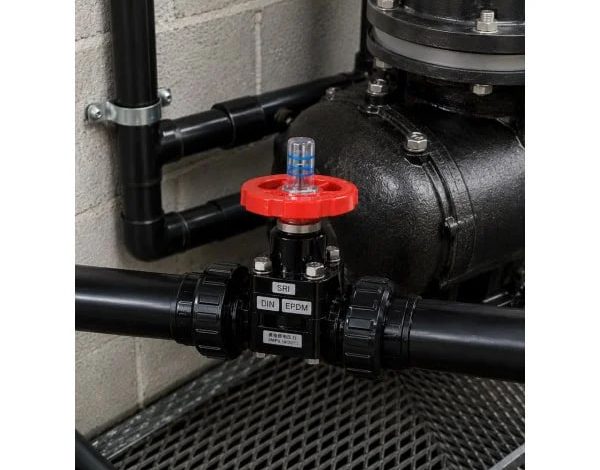 Industrial shut-off valve with a red handwheel on a black pipe network beside a large pump and concrete wall.