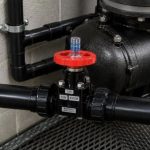 Industrial shut-off valve with a red handwheel on a black pipe network beside a large pump and concrete wall.